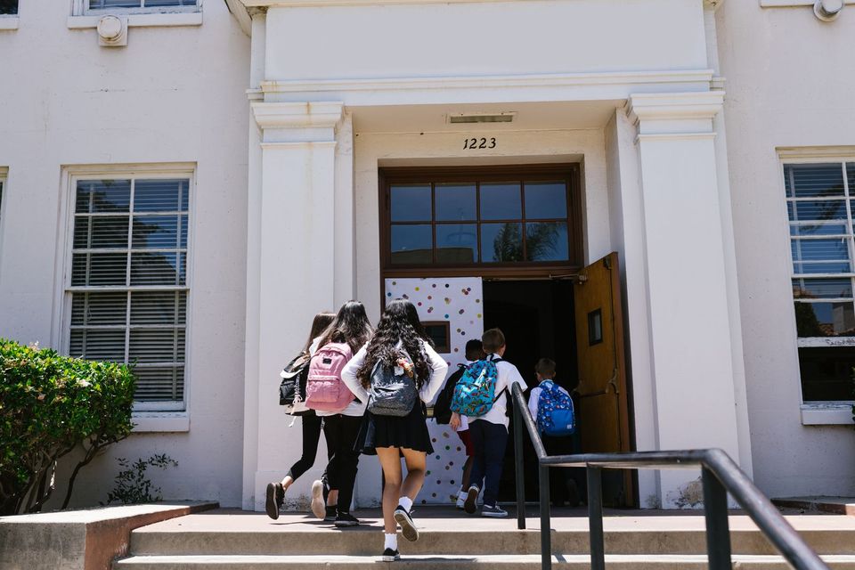 Children with backpacks walking up the steps into a school, seen from behind